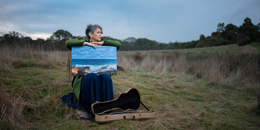 A woman with short, gun-metal grey hair sits on a low rock in a field of long grass with gum trees visible in the distance behind her. She is wearing a long, dark blue dress and a light forest green jacket. She is looking to her left and has her forearms resting on the top edge of a painting of a shoreline with crashing blue waves. The painting rests on her lap facing the viewer.