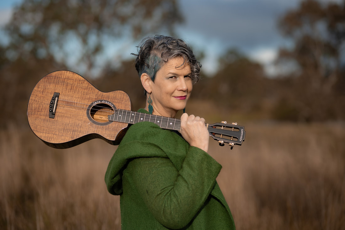 A woman with short, gun-metal grey hair stands side on with her head turned to look at the camera. She holds a baritone ukulele by its neck, resting it on her shoulder. She is wearing a light forest green hooded jacket with the hood down and stands in a clearing of brown long grass with gum trees behind her. 