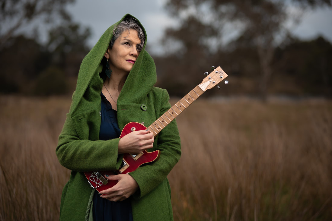 A woman with short, gun-metal grey hair stands facing the camera and looking up at the sky with her head tilted slightly to her left. She cradles a red electric baritone ukulele in her left arm with the fingers of her right hand resting on the strings where the neck joins the body of the uke. She is wearing a light forest green hooded jacket with the hood up and partially covering her left cheek and stands in a clearing of brown long grass with gum trees behind her. 