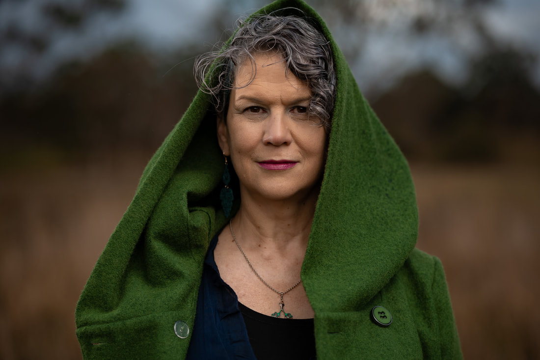 A woman with short, curly, gun-metal grey hair faces the camera. The photo is a close portrait with only her head and shoulders visible. She looks straight at the camera with a slight smile. She is wearing a dark blue dress and a light forest green hooded jacket with the large hood up and the jacket unbuttoned. The background is a blur of grey and brown suggesting an open field with trees behind..