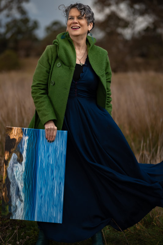 A woman with short, curly, gun-metal grey hair stands face on with her head tilted up slightly as she laughs. She holds a painting of a shoreline with blue crashing waves in her right hand so that it hangs beside her leg. She is wearing a long, dark blue dress, a silver celtic pendant, teal drop earrings and a light forest green hooded jacket with the hood down and stands in a clearing of brown long grass. The skirt of her dress is billowing out the front of the jacket in the wind and there are gum trees behind her but out of focus.. 